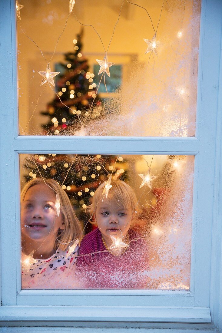 Geschwister am Fenster mit Lichterkette und aufgesprühtem Kunstschnee