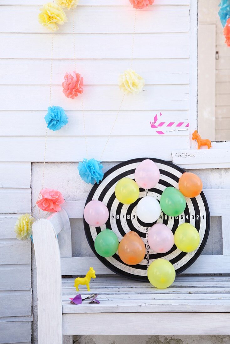 Dartscheibe mit bunten Luftballons vor weißer Holzwand mit Papierblumen-Girlande