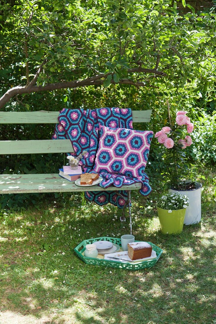 Crocheted cushions and blanket on garden bench and tray of cake on lawn