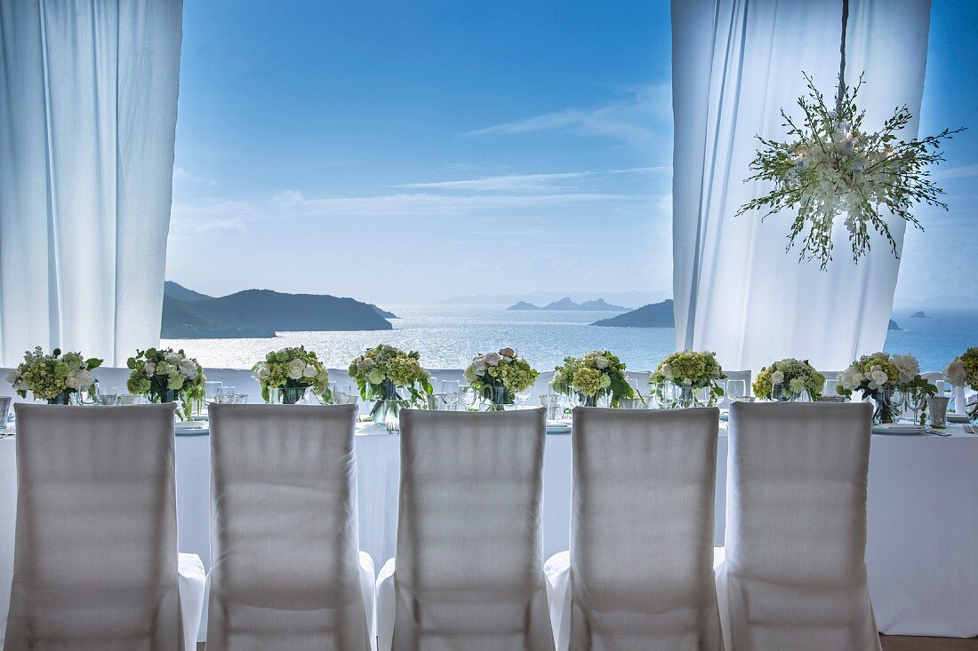 Chairs with white loose covers around festively set wedding dinner table with sea view