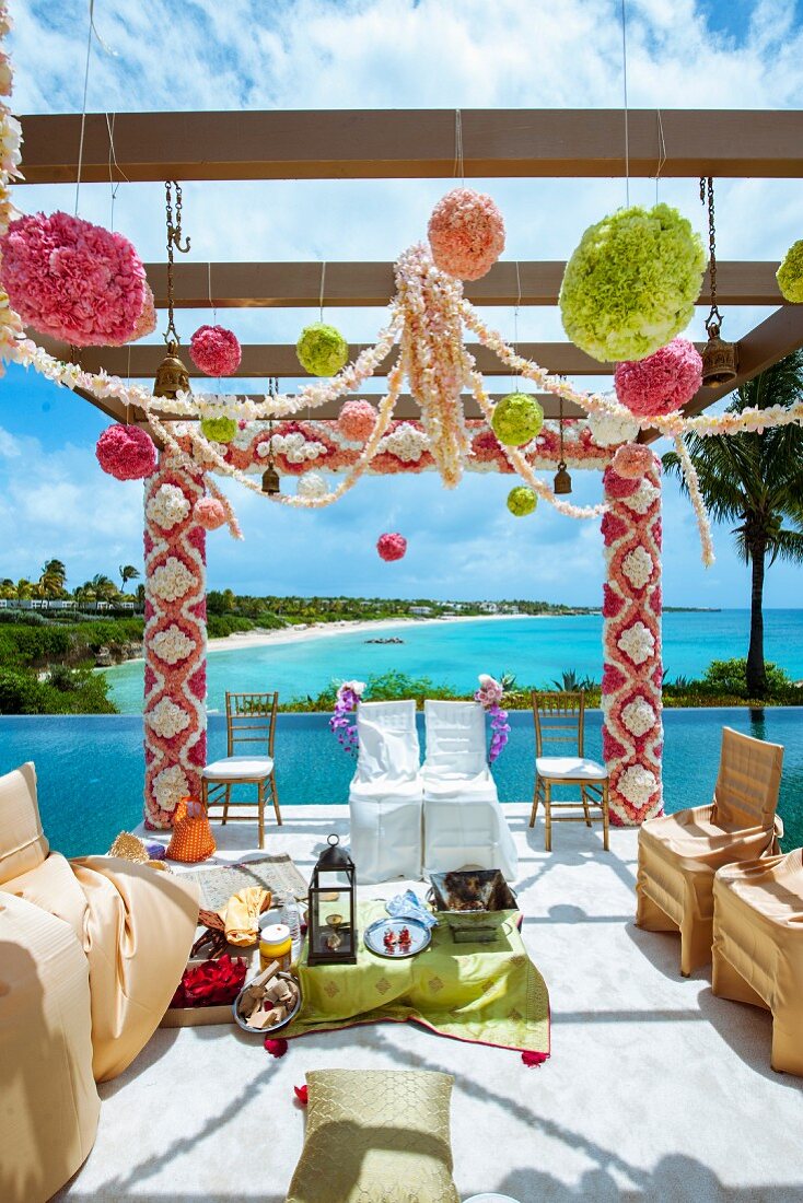 Indian wedding - chairs with white and gold loose covers below pergola decorated with flowers next to ocean