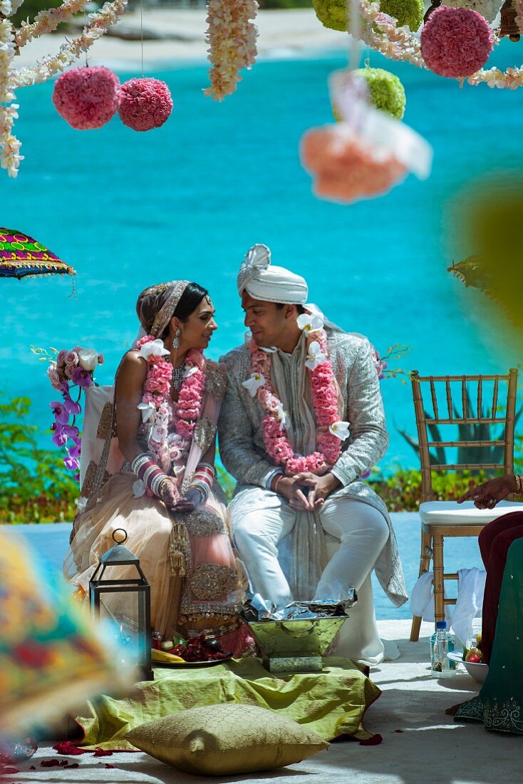 Indian bride and groom adorned with pink flower garlands against backdrop of turquoise sea