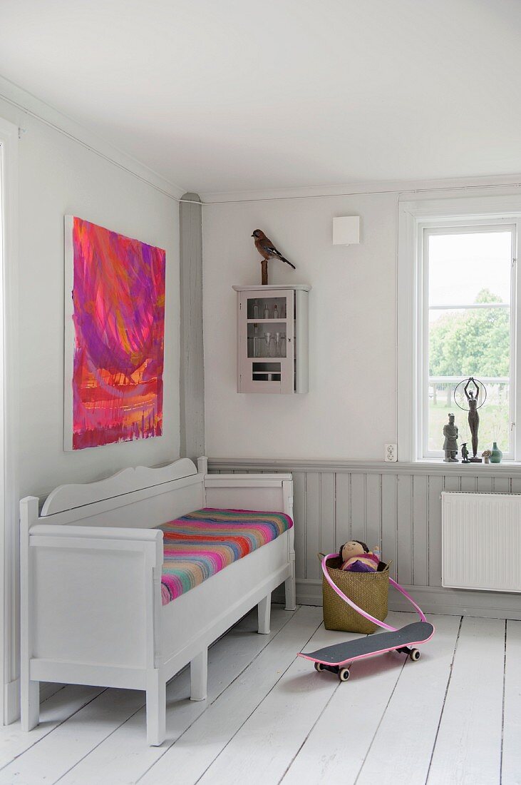 Rustic white bench with brightly coloured seat cushion below modern artwork in corner
