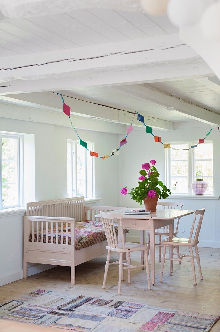 Pink-painted chairs, table and bench below windows and colourful garland in corner of rustic room