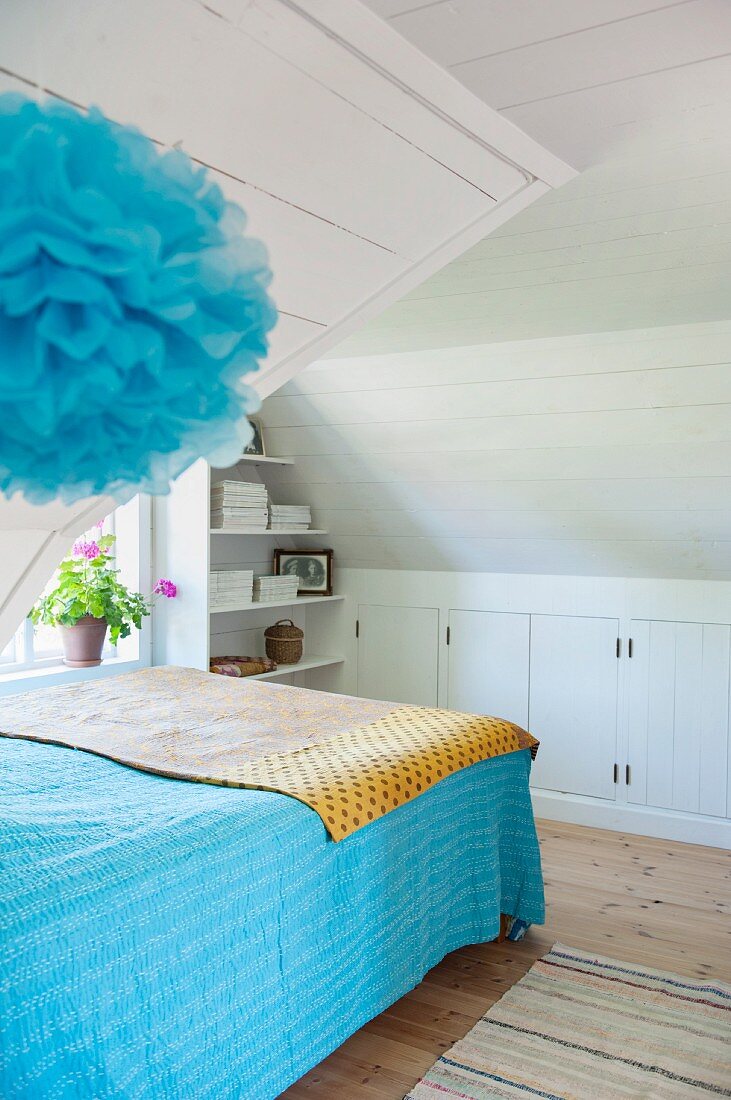 Bed with blue bedspread in white wood-clad attic room with blue pompom in foreground