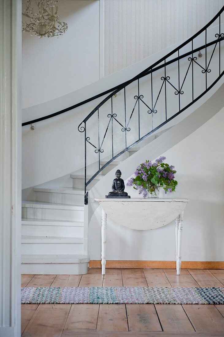 Vase of lilac and Buddha figurine on white wooden table at foot of winding staircase with delicate wrought iron balustrade in foyer