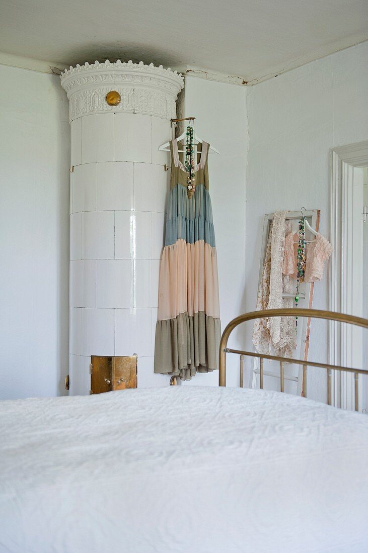 Blick über Bett auf antiken, runden Kachelofen mit weissen Fliesen und daran aufgehängtem Sommerkleid