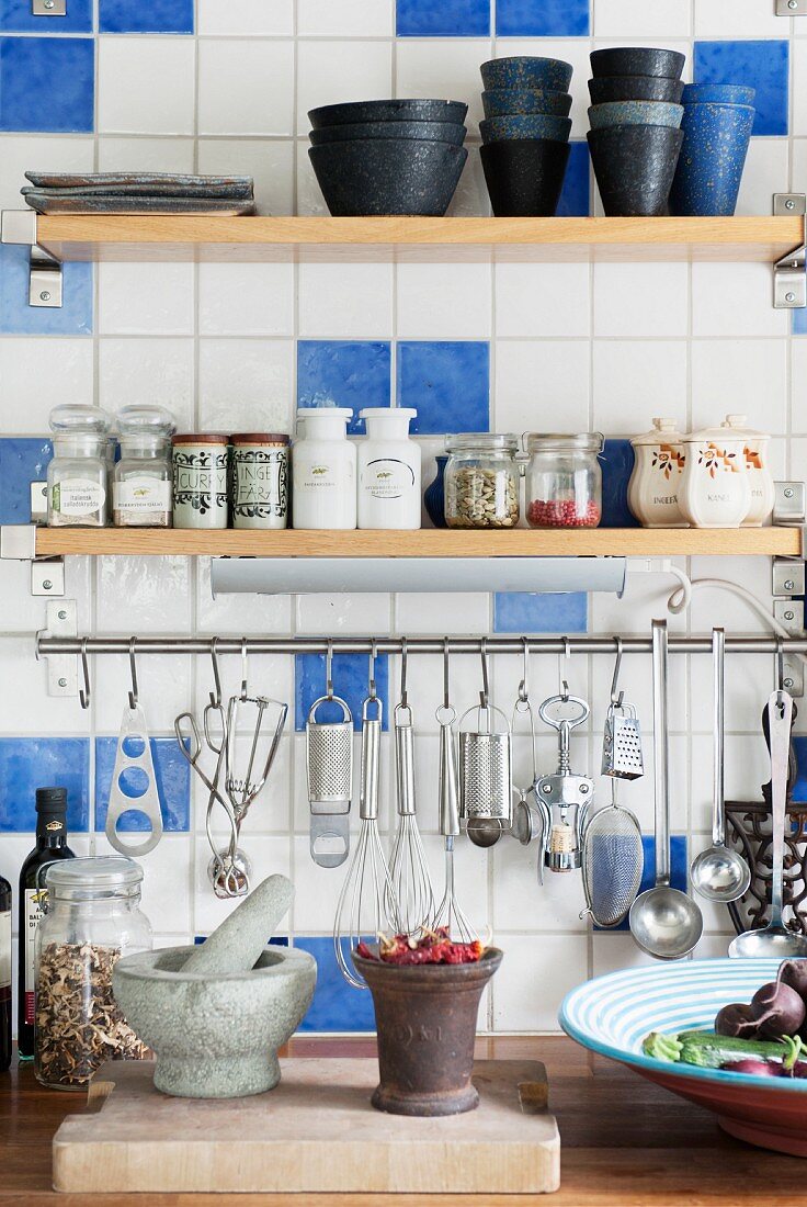 Weiß-blau geflieste Küchenwand, mit Reglabrettern für Gewürze und Geschirr über Edelstahlstange mit aufgehängten Kochutensilien
