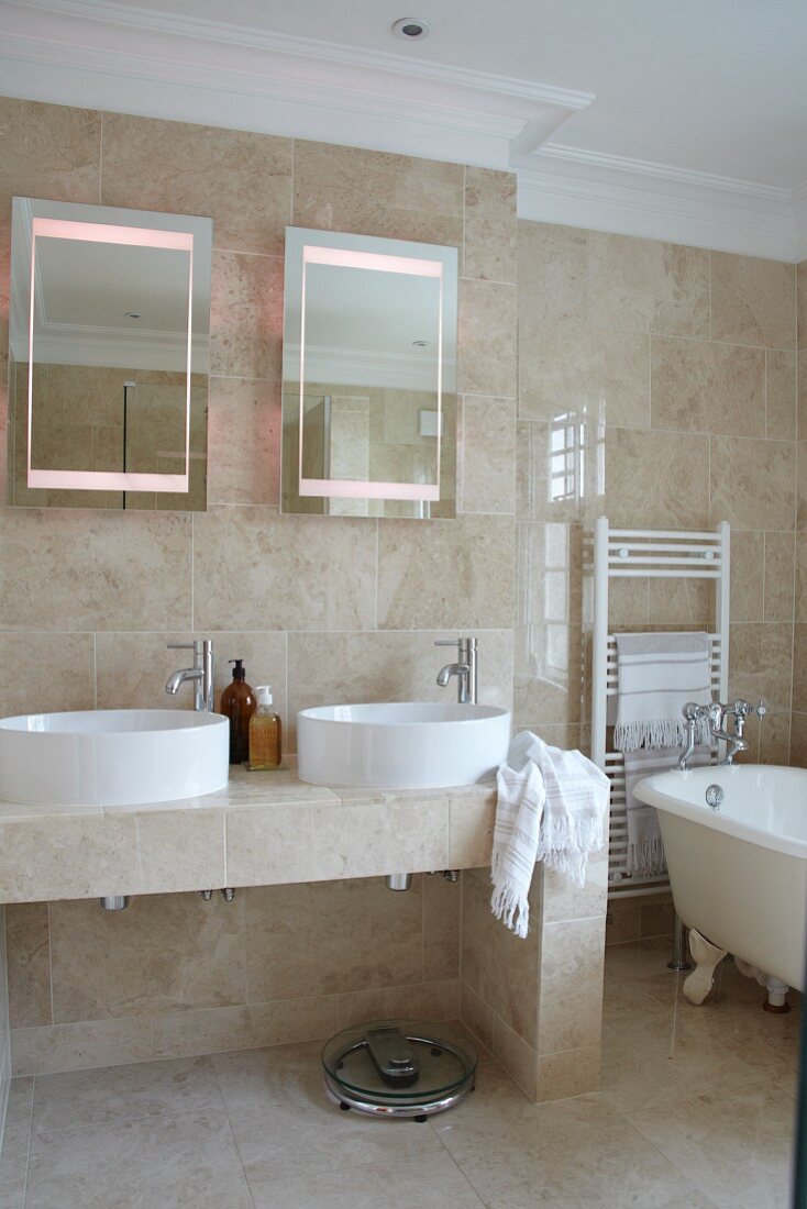 Bathroom with beige stone tiles, twin countertop sinks, two mirrors and free-standing, vintage-style bathtub