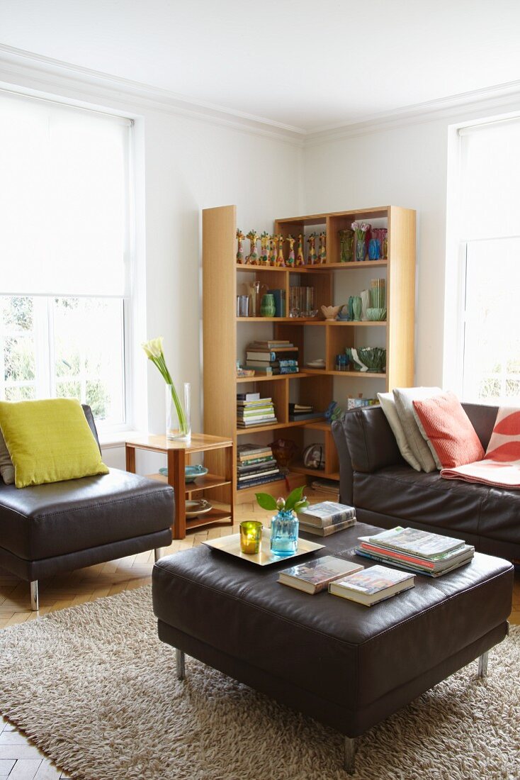 Gemütliches Wohnzimmer mit dunkelbraunen Ledermöbeln, Teppich und Holz-Eckregal zwischen hellen Sprossenfenstern