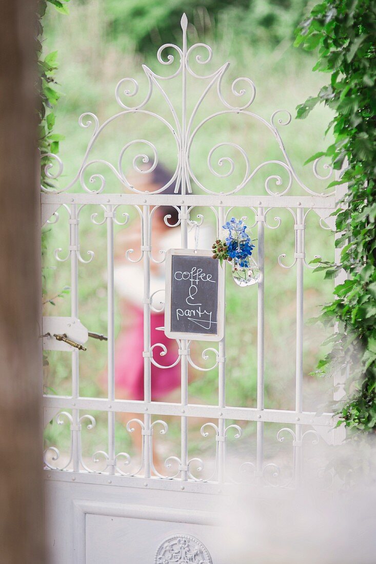Schiefertafel mit Kaffeeeinladung und Blütendeko an romantischer, schmiedeeiserner Gartentür