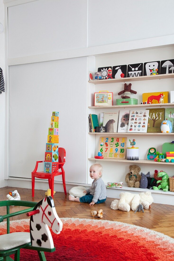 Toddler sitting on parquet floor below shelves of toys; retro rocking horse in foreground