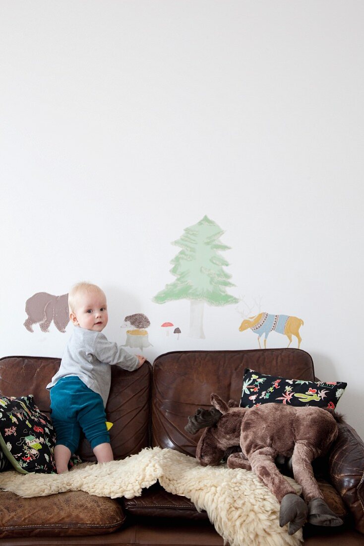 Toddler standing on sheepskin rug on brown leather sofa below animal wall-stickers