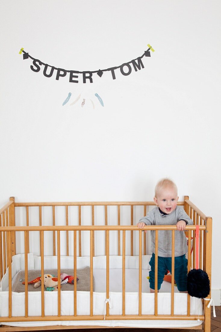 Kleinkind im Gitterbett stehend, an Wand Jungenname aus schwarzen Buchstaben aufgehängt