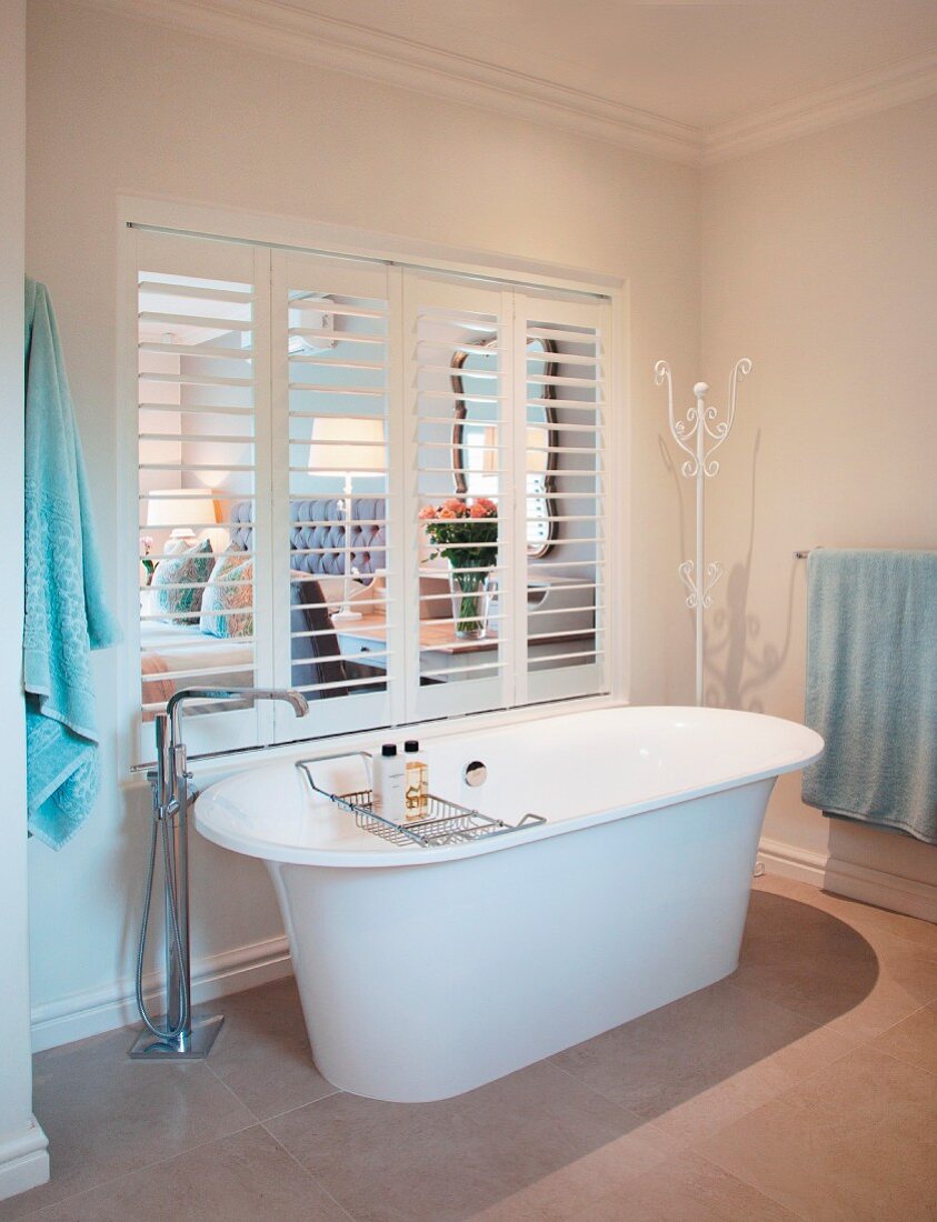 Freistehende Badewanne mit Standarmatur vor Innenfenster mit offenen Lamellen Fensterläden und Blick ins Schlafzimmer