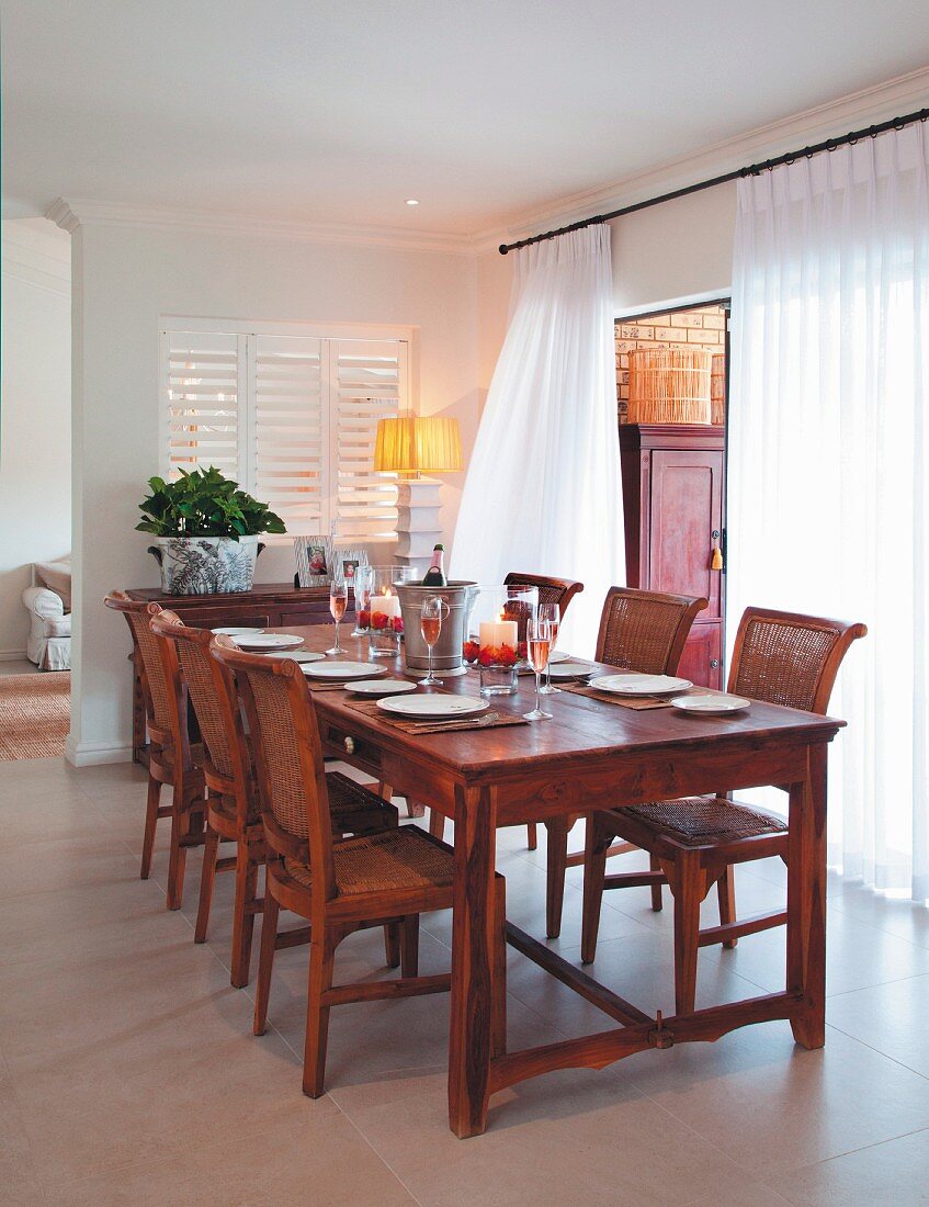 Gedeckter Esstisch und passende Stühle aus rötlichem Holz, vor Fenster mit bodenlangem, transparentem Vorhang