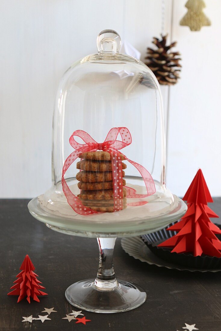 Gestapelte Weihnachtsplätzchen mit Schleife auf Etagere unter Glasglocke, seitlich Weihnachtsbäumchen aus rotem Papier