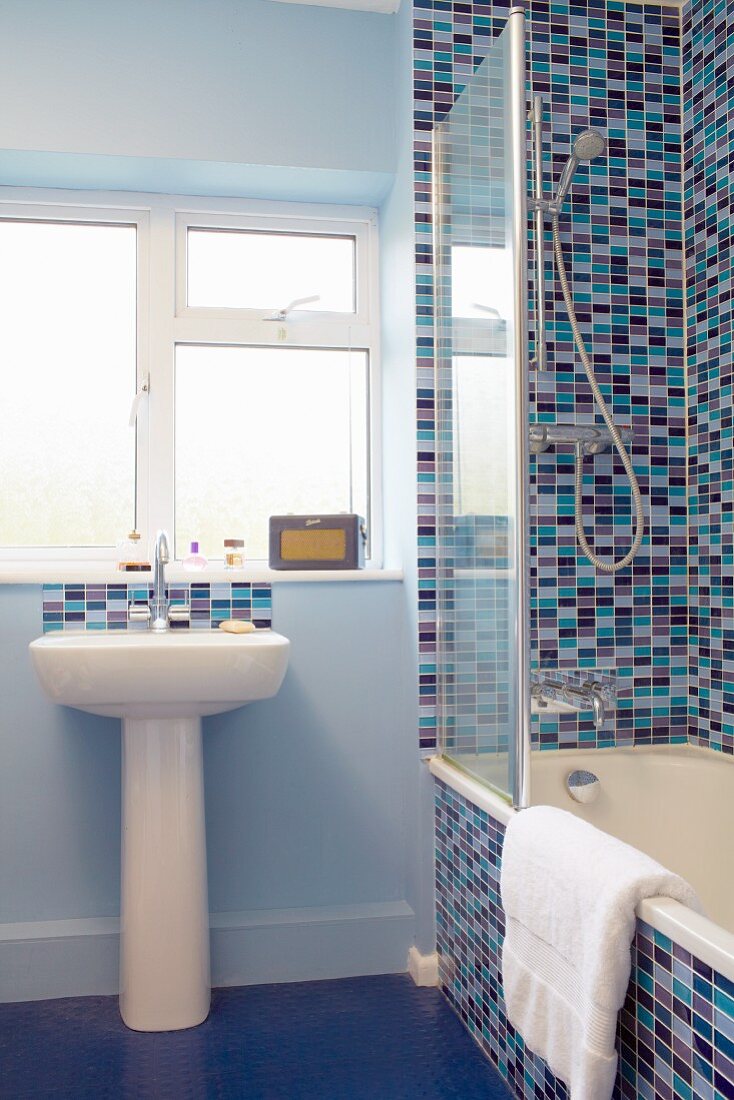 Pedestal sink below window next to bathtub against wall tiled in various shades of blue and red