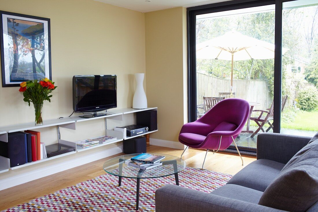 Wohnraumecke, runder Glas Tisch und lila Sessel vor Terrassentür, seitlich offenes Lowboard an pastellgelber Wand