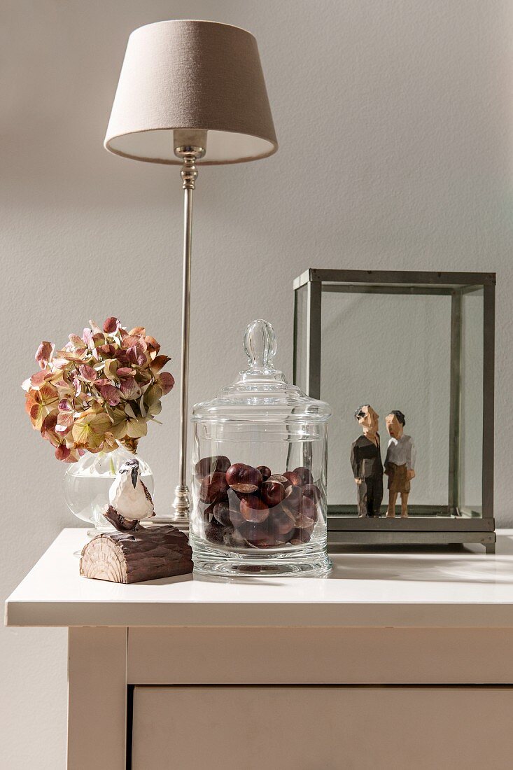 Mit Kastanien gefüllte Glasdose vor Tischleuchte und Hortensienblüte in Vase, daneben Holzskulpturen in Glasvitine