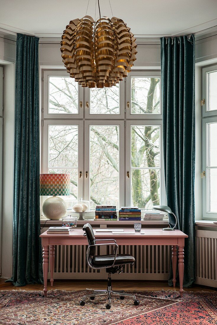 Rosa lackierter Schreibtisch vor Erkerfenster mit bodenlangen Vorhängen, im Vordergrund Pendelleuchte mit glänzendem Lampenschirm