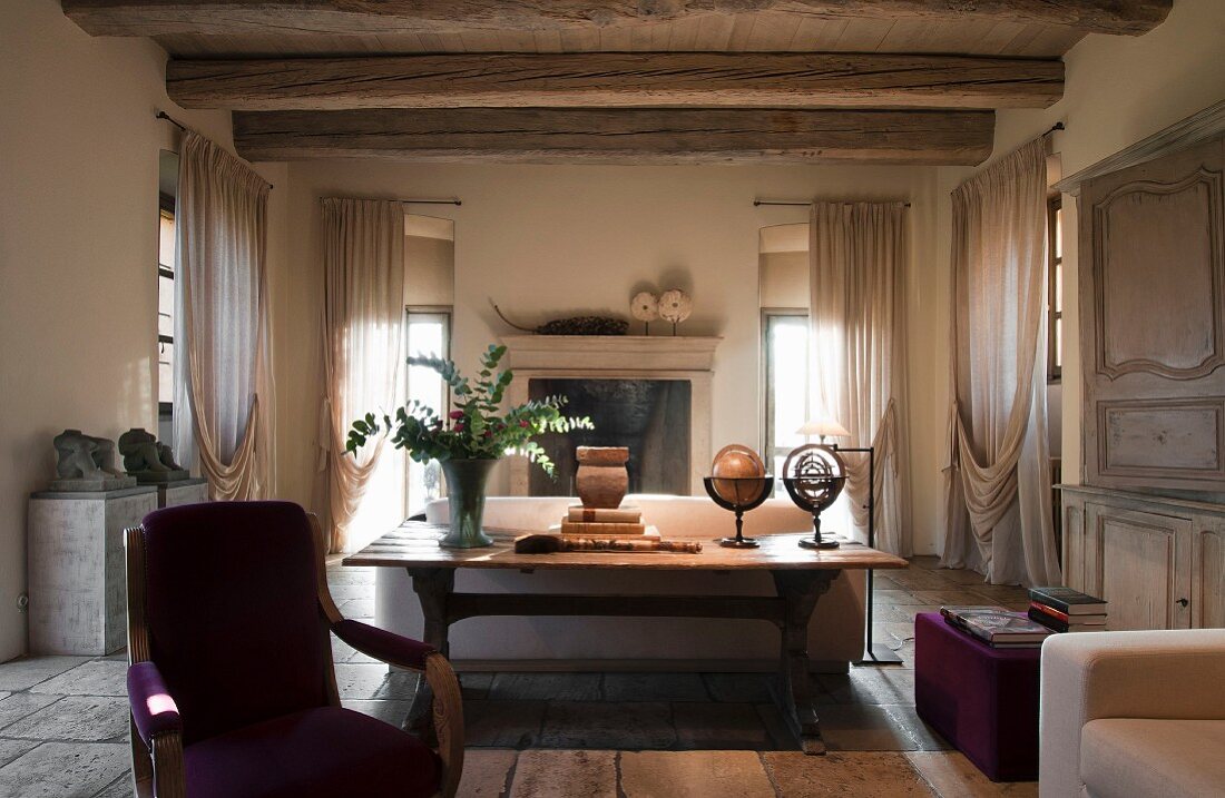 Wohnraum einer alten, renovierten Villa, rustikaler Holztisch vor Rückseite eines Sofas, an Fenstern drapierte, bodenlange Vorhänge