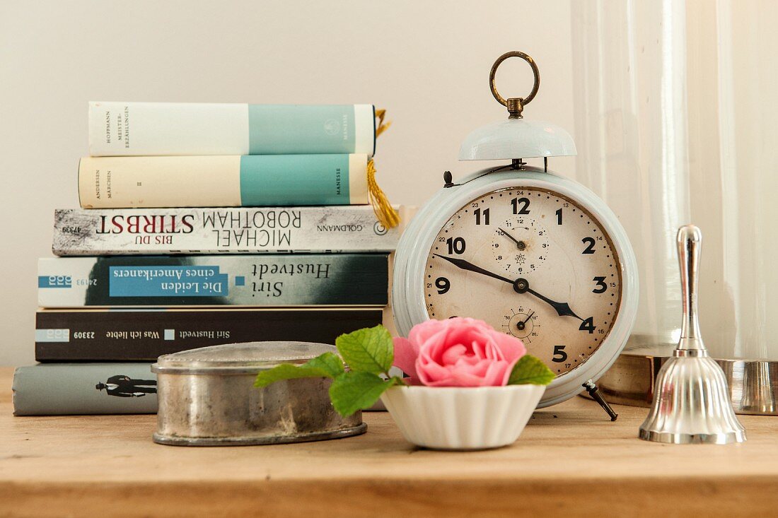 Silver tin and rose in pot in front of stacked books and vintage-style alarm clock