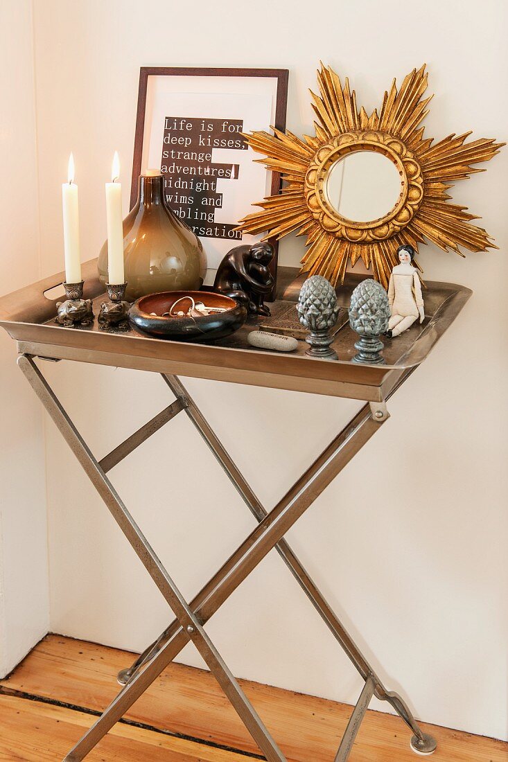 Tabletttisch aus Metall mit brennenden Kerzen und Dekoobjekten, Goldrahmenspiegel in Strahlenform