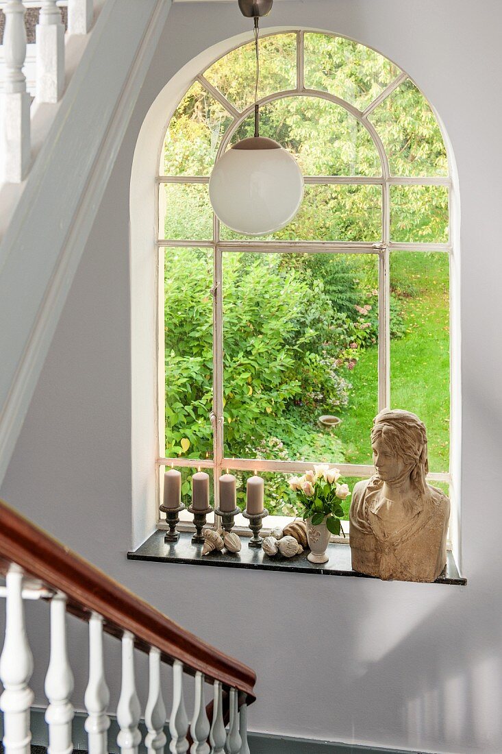 Rundbogenfenster mit Gartenblick im Treppenhaus, Frauenbüste und Kerzenständer auf Fensterbank