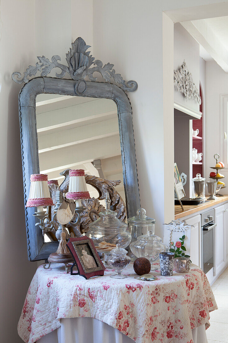 Vintage ornaments and glass jars in front of mirror with ornate papier mâché frame on side table with floral tablecloth