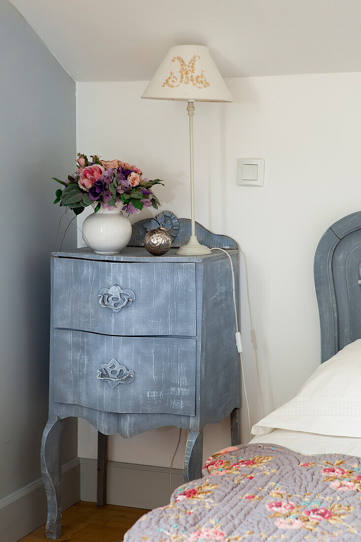 Vase of flowers and embroidered table lamp on blue-grey bedside cabinet decorated with corrugated carboard