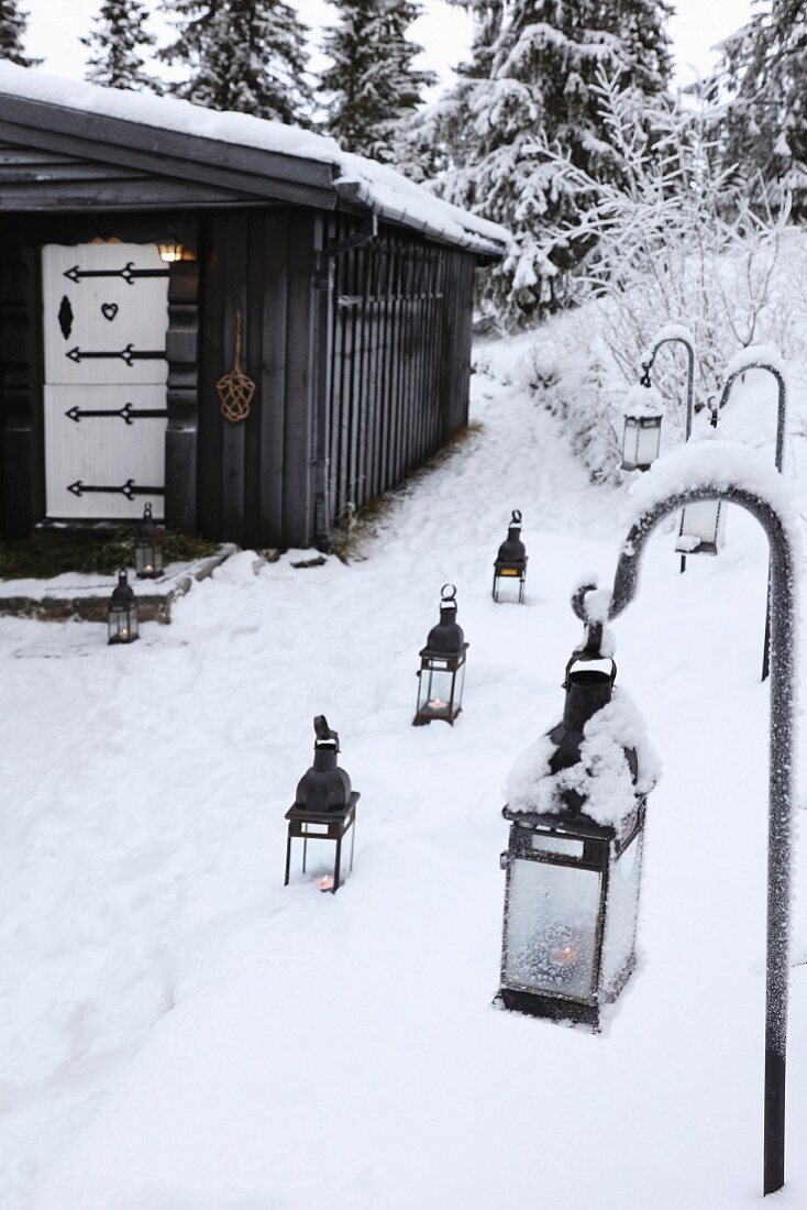Laternen im Schnee vor einer schwarzen Holzhütte