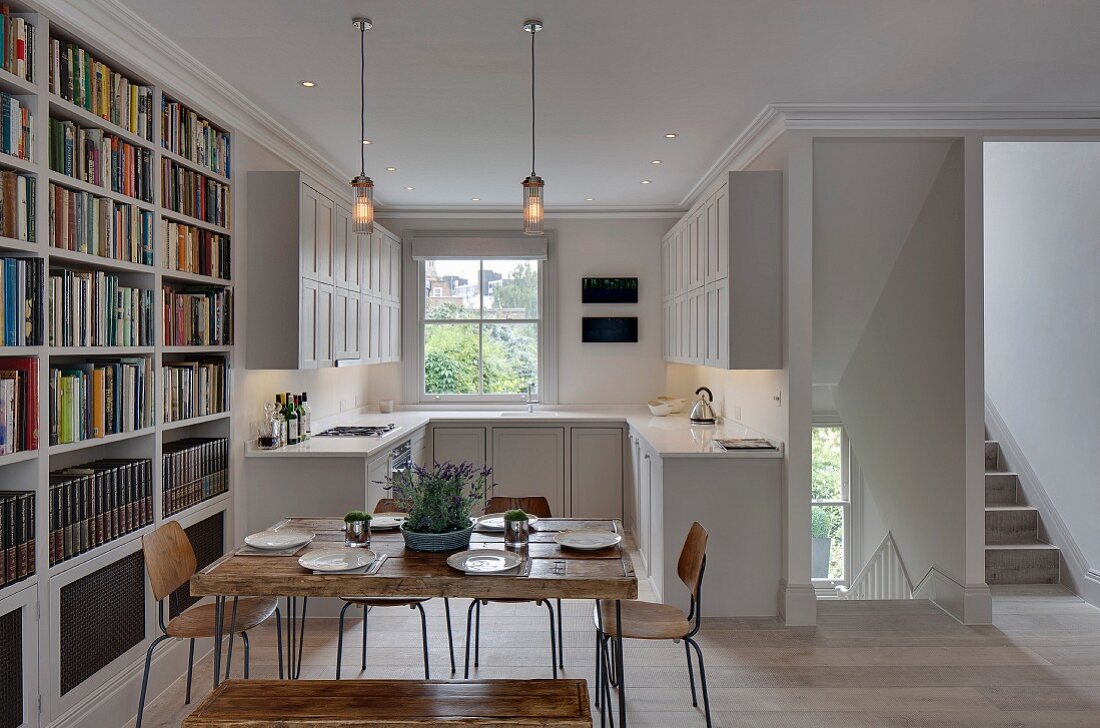 Square, rustic wooden table, vintage chairs and bookcase in front of white fitted kitchen with panelled fronts