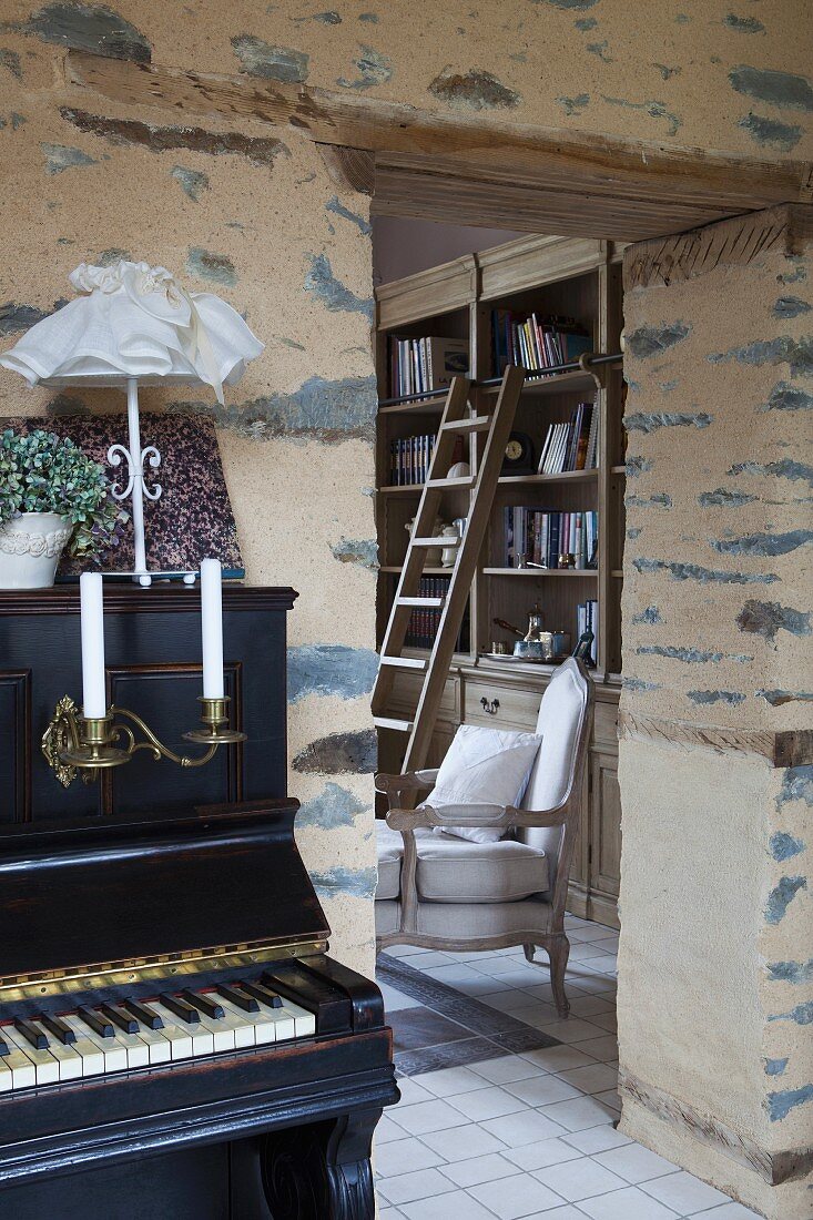 Piano in front of rustic wall with view of reading chair in library through open doorway