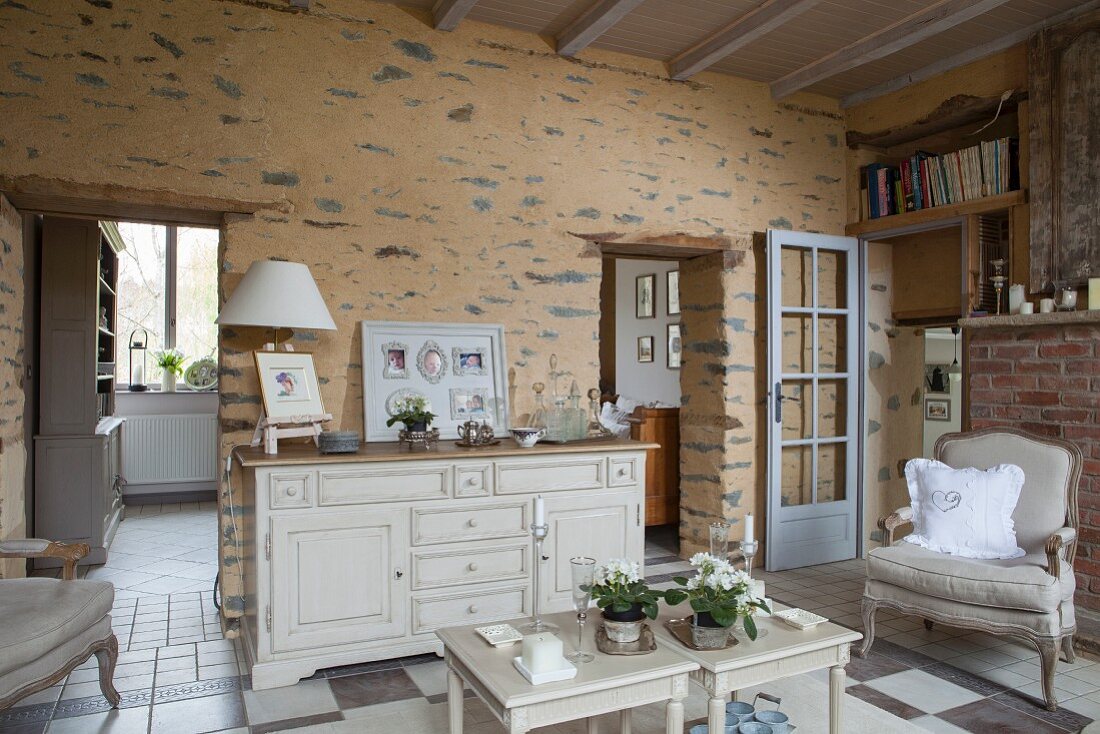 Ländliches Wohnzimmer mit nostalgischem Flair; Beistelltische und weisses Sideboard vor rustikal gestalteter Wand