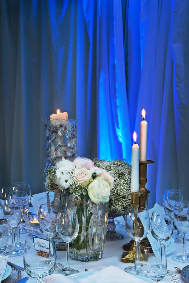 Kerzenhalter aus Messing mit brennenden Kerzen auf festlich gedeckten Tisch, vor blau angestrahltem Vorhang im Hintergrund