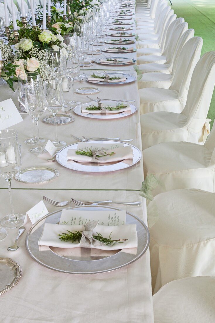Long festive table with decorated place settings, silver plates, chairs with white covers in front of it