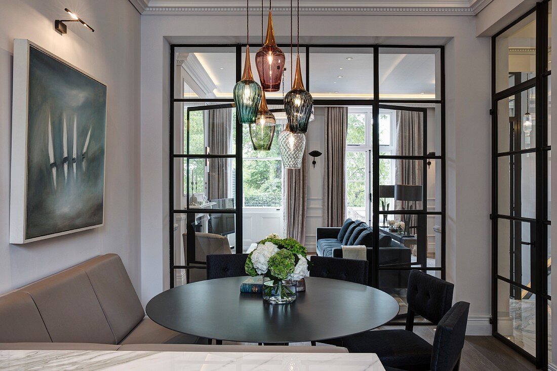 Upholstered bench, black chairs and round table below pendant lamps with glass lampshades of various colours in front of glass partition wall