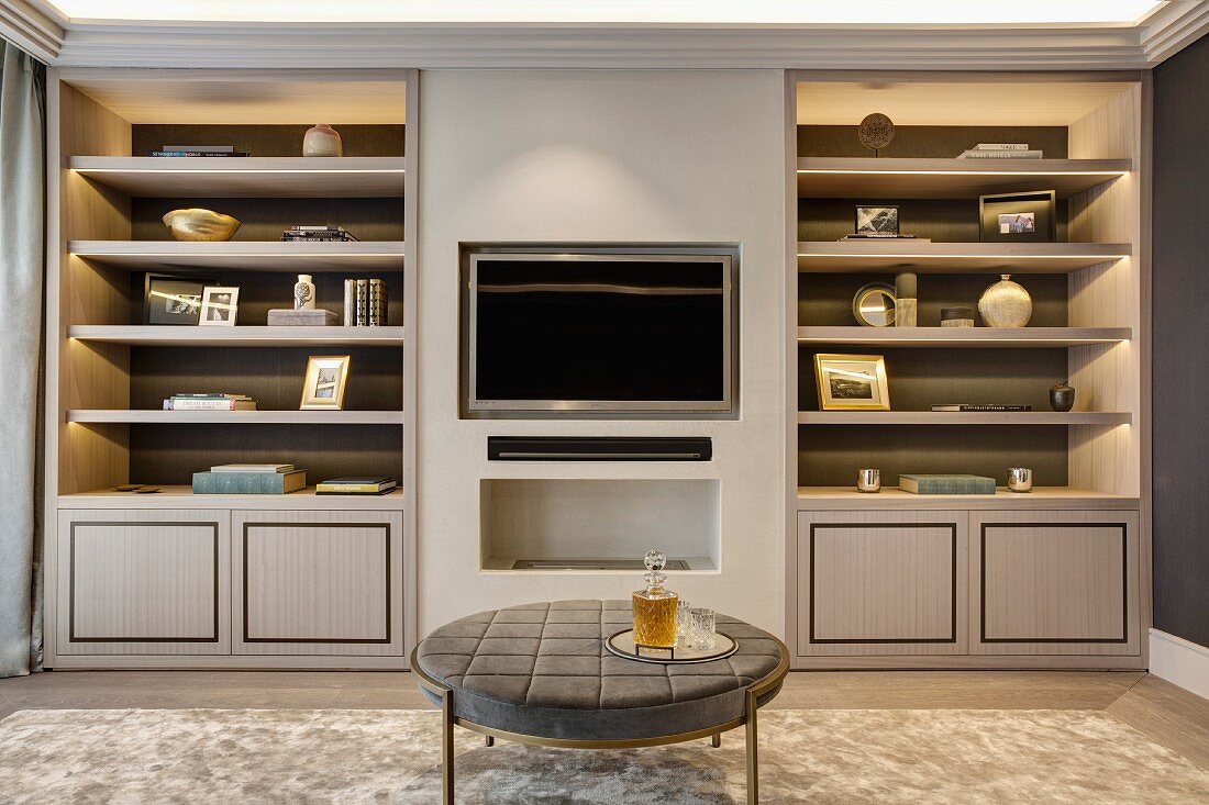 Round table in front of flatscreen TV recessed in wall between fitted cupboards and shelves