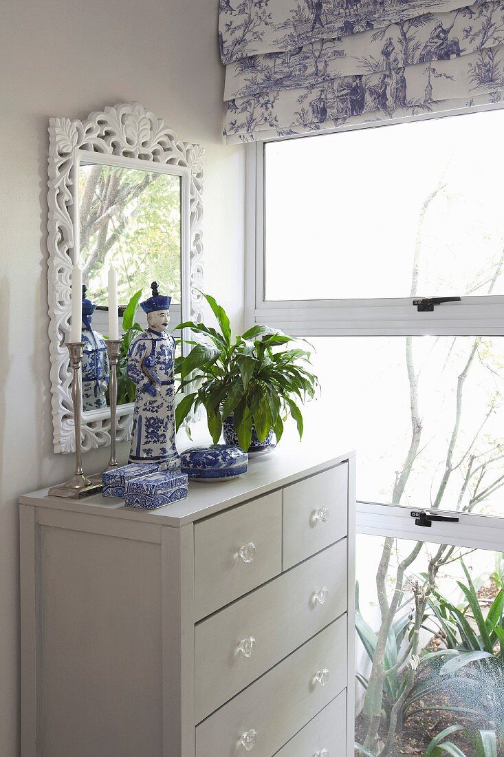Chinese porcelain on chest of drawers and mirror with floral frame in front of window with toile de jouy Roman blind