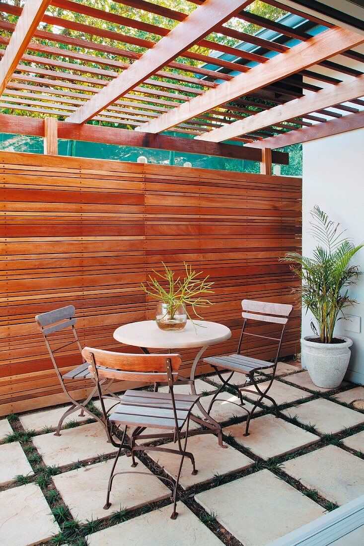Bistrotisch mit Stühlen auf Terrasse mit Rasenfugen im Plattenbelag, Holzwand mit Pergola