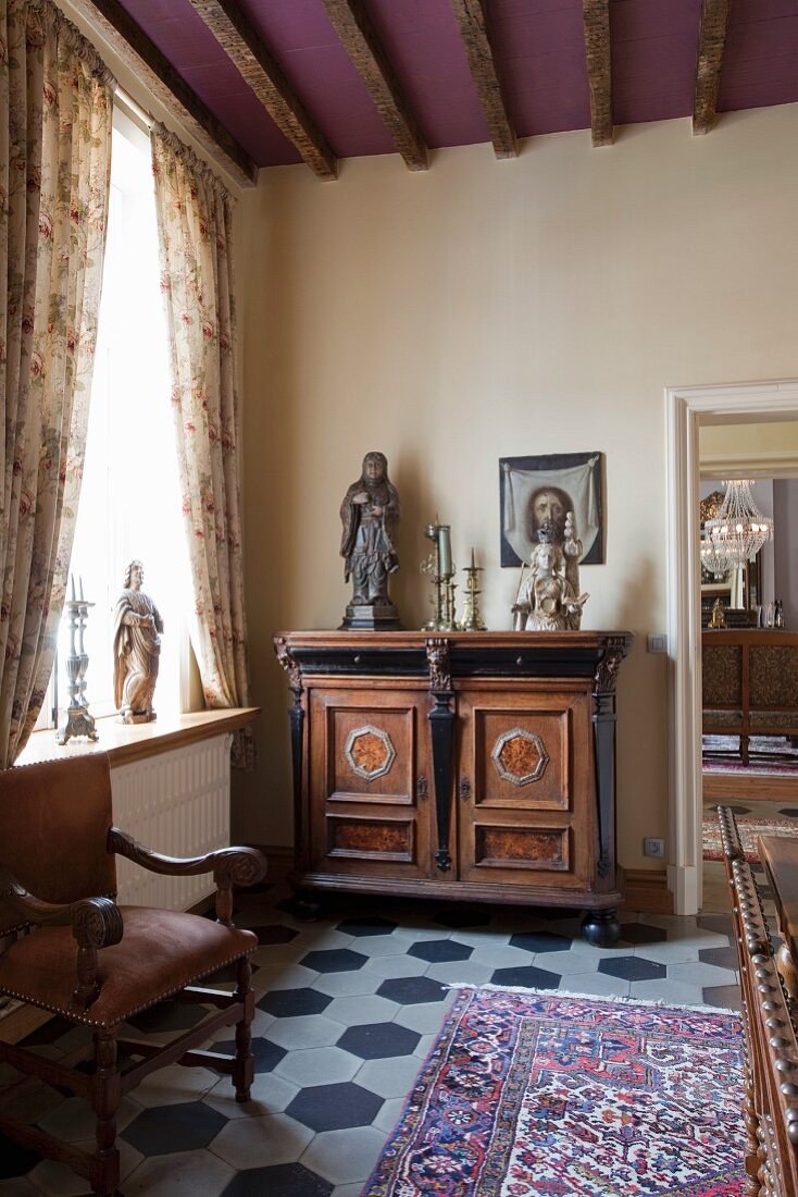Sculptures and candlesticks on top of antique, elegant cabinet next to window with draped curtains