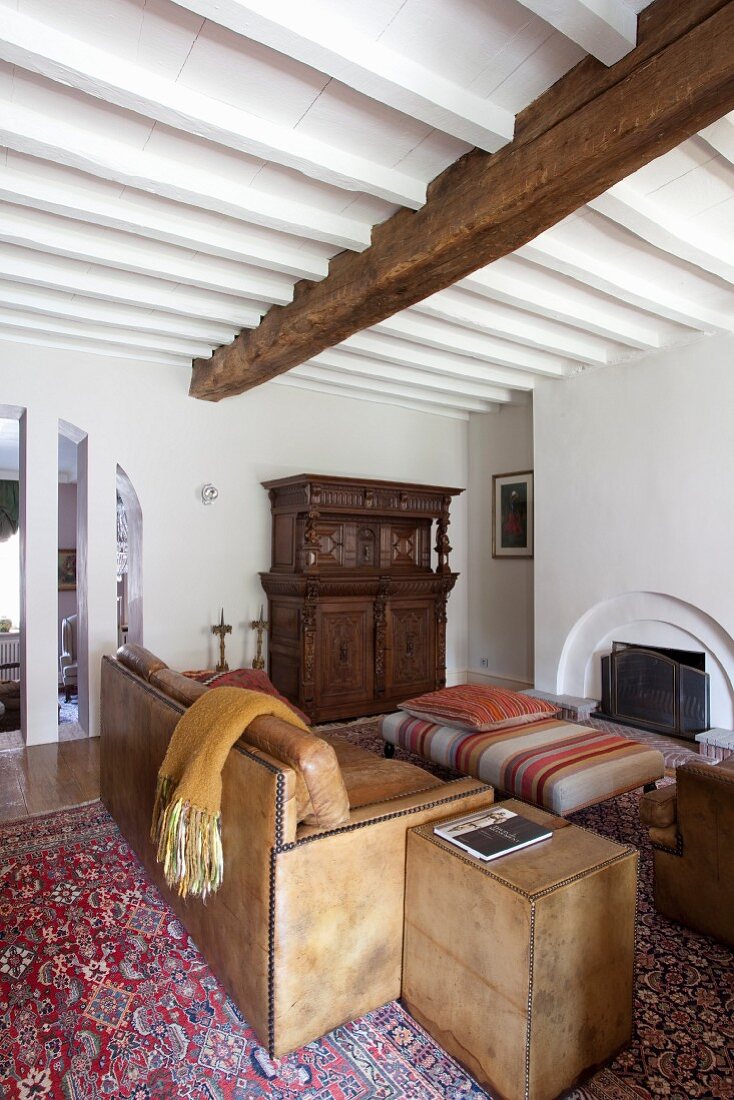 Traditionelles Wohnzimmer mit hellbrauner Ledercouch, Beistelltisch und Polstertisch vor offenem Kamin, weiße Holzbalkendecke
