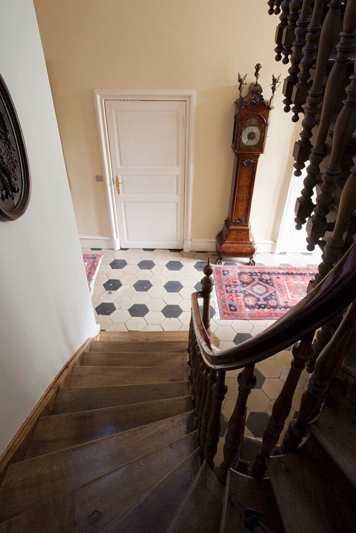 View down winding wooden staircase into hallway with honeycomb floor tiles and antique long-case clock