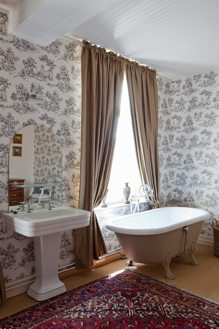 Free-standing bathtub below window with light brown draped curtains next to vintage pedestal sink in traditional ensuite bathroom