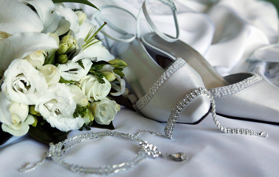 weiße Hochzeitsschuhe mit Schmuckstücken neben weißem Rosenstrauss auf Satintuch