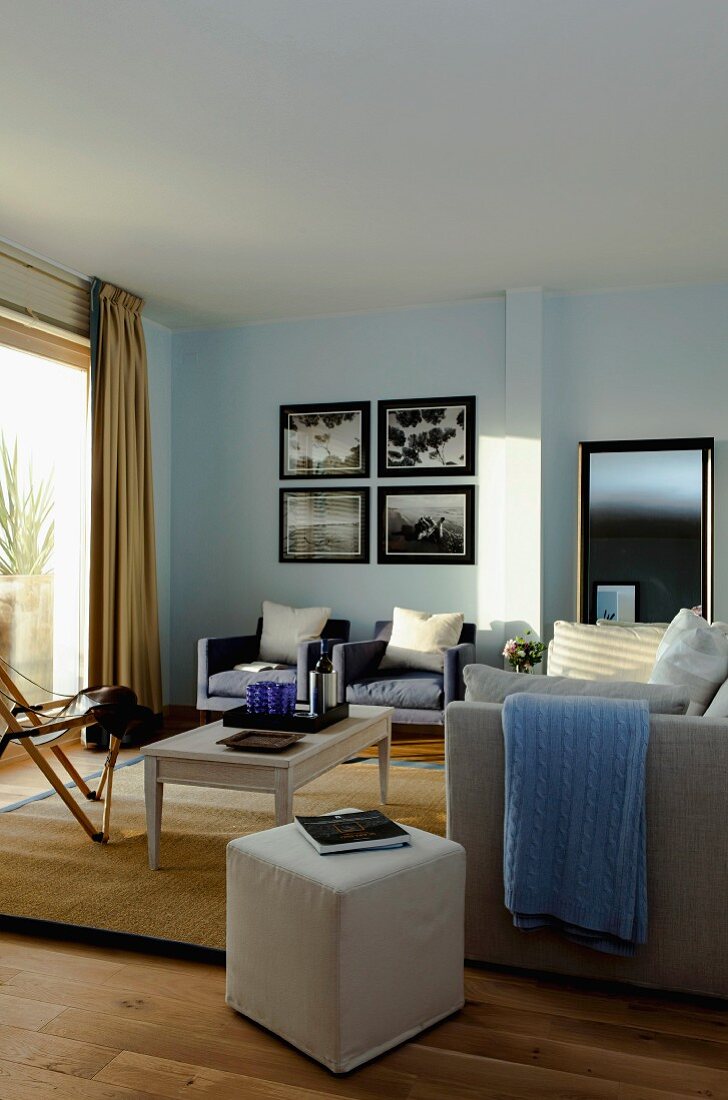 Wohnzimmer mit verschiedenen Sitzmöbeln, Hocker mit hellem Bezug, im Hintergrund Sessel an hellblau getönter Wand unter Bildersammlung