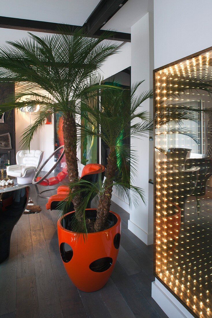 Loungebereich mit Zimmerpalme in orangerotem Übertopf und leuchtendes Kunstobjekt an Wand, Retroflair