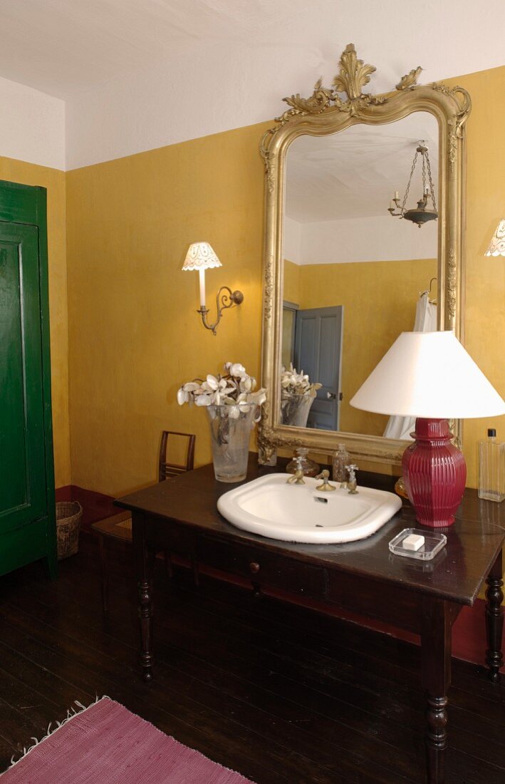 Washstand made from desk with integrated sink and table lamp in front of elegant, gilt-framed mirror on yellow-painted wall