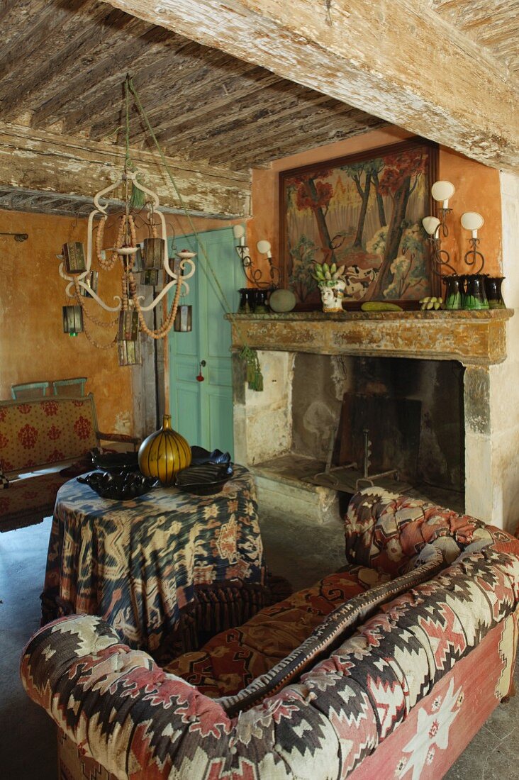 Sofa mit folkloristisch gemustertem Bezug und runder Tisch mit Tischtuch, seitlich offener Kamin in rustikalem Ambiente